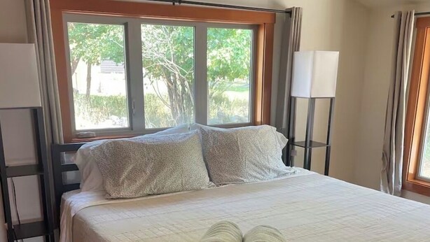 A neatly made bed with rolled towels, a bedside table, a lamp, and a window with a view of greenery. - Cozy Rattlesnake Creekside Cabin Retreat  (Missoula)