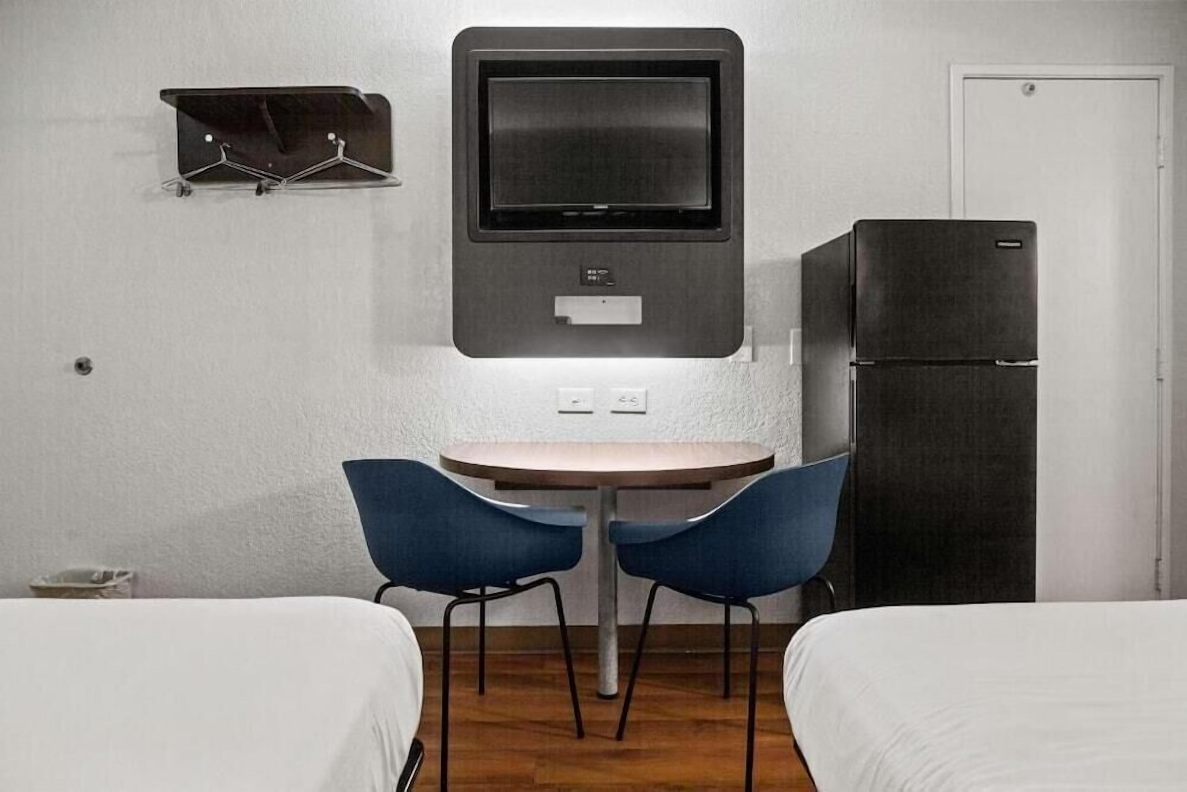 A hotel room with a bed, a desk with a computer monitor, a refrigerator, and two chairs.