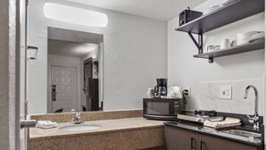 Un baño moderno con encimera de granito, lavabo, espejo, cafetera y repisa con artículos de aseo.