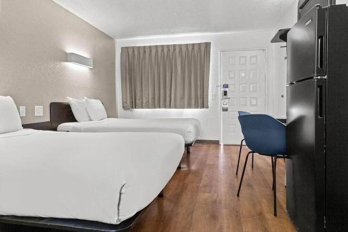 Ein Hotelzimmer mit zwei Betten, einem Schreibtisch, einem Stuhl, einem Kühlschrank und einem Fenster mit Vorhängen.