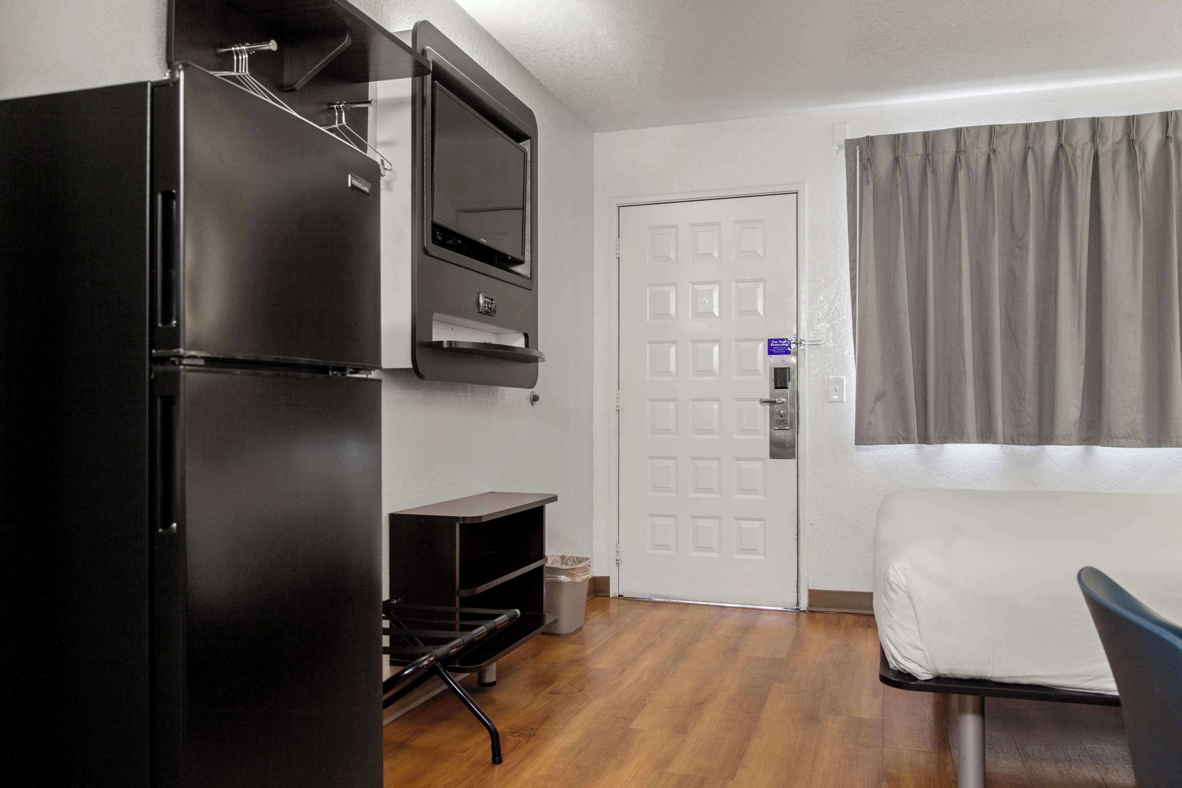 A compact hotel room with a kitchenette, a bed, a TV, and a window with curtains.