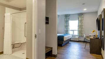 Ett modernt hotellrum med en duschhörna, en säng med ett blått överkast, ett skrivbord med en lampa och ett fönster med mönstrade gardiner.