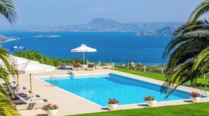 Relax by the pool with a view of the sea and distant mountains, surrounded by lounge chairs and lush greenery.