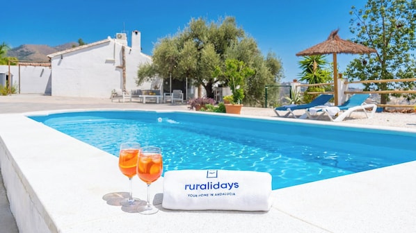 Ein Poolbereich mit Liegestühlen, einem Reetdach-Sonnenschirm und einem Schild mit der Aufschrift 'ruraldays IHR ZUHAUSE IN ANDALUSIEN'.