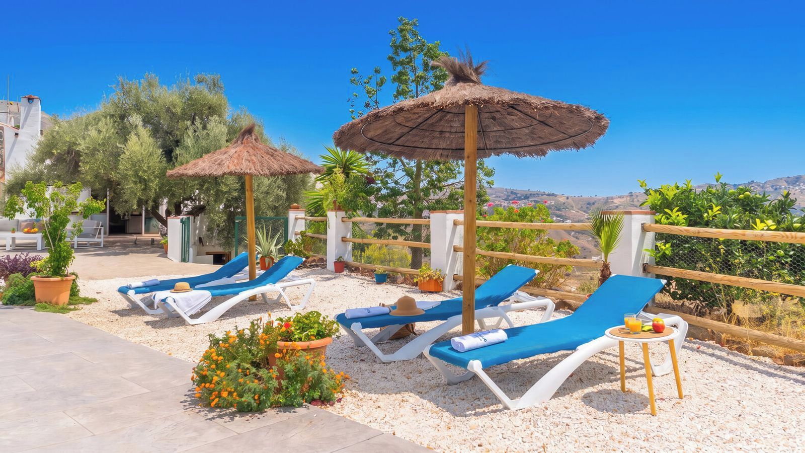 Eine sonnendurchflutete Lounge-Bereich im Freien mit mehreren blauen Liegestühlen, einige mit Sonnenschirmen, umgeben von Kübelpflanzen und einem Kiesweg.