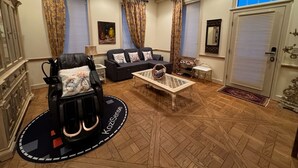 A living room with a black leather chair, a sofa, a coffee table, and a ceiling fan. There are curtains on the windows and a door with a mirror. - Chateau Herbe - Shakespeare 1 Bedroom Suite w/ Kitchen (Ashland)