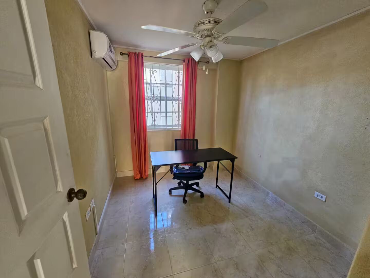 Una habitación con ventilador de techo, ventana con cortinas rojas, una mesa con una silla y piso de baldosas.