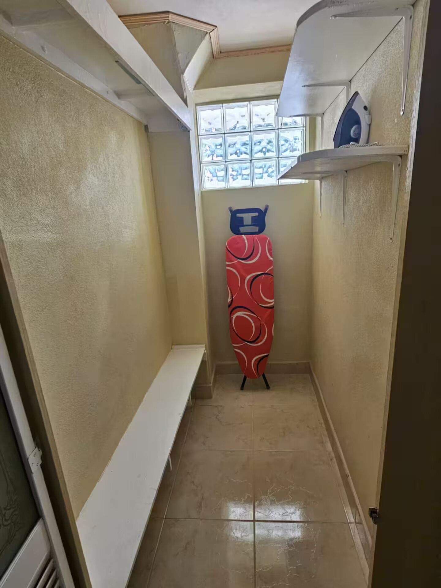 Un couloir étroit, avec un banc sur la gauche, une fenêtre à carreaux et, sur la droite, une planche à repasser rouge dotée d’un manche bleu.