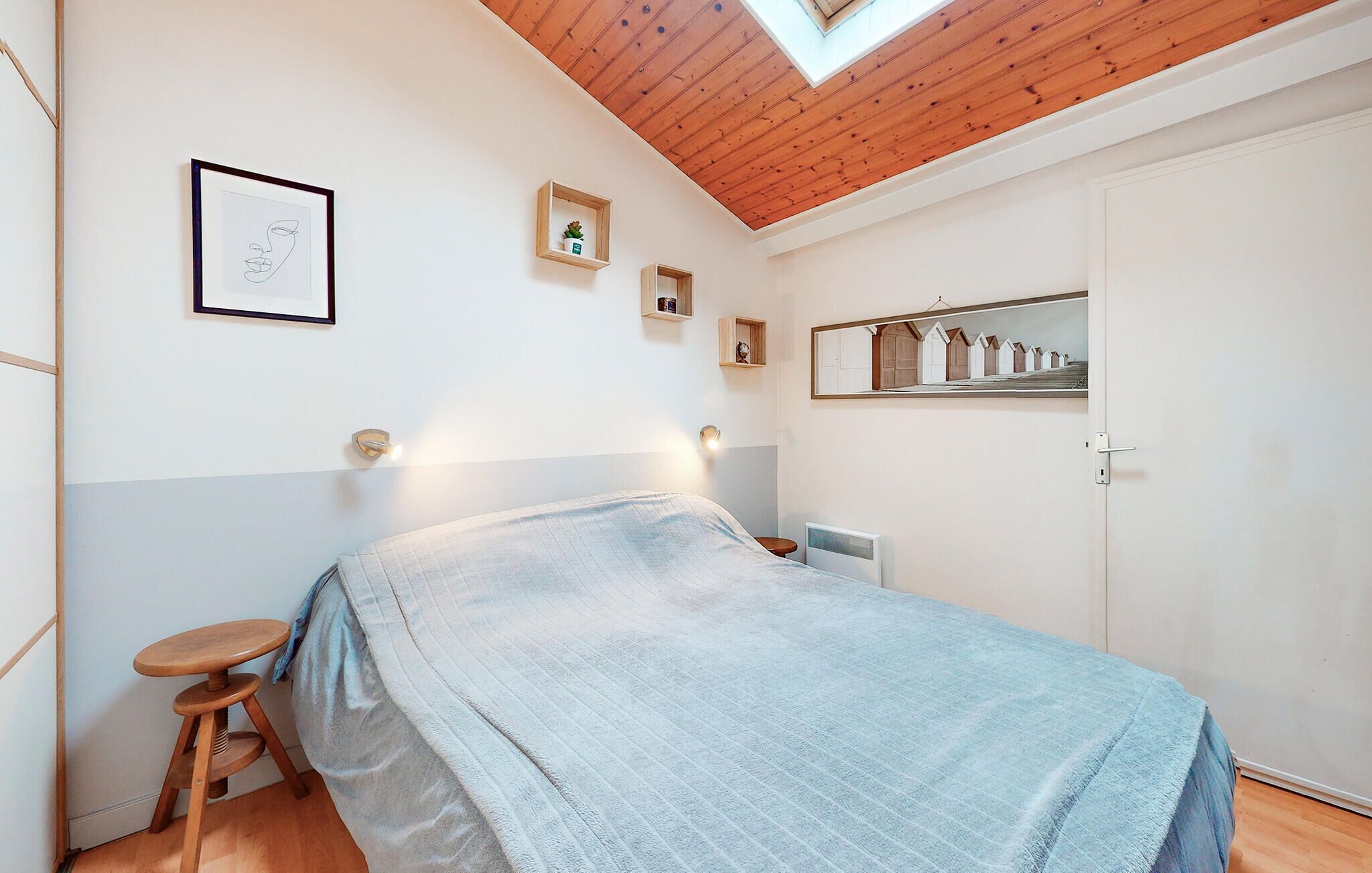 Une chambre à coucher avec un lit, un tabouret en bois, une étagère ornée d’objets décoratifs et une lucarne.