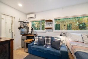Un espace de vie compact comprenant un coin cuisine, un canapé avec des coussins, un lit et de grandes fenêtres donnant sur la forêt.