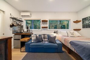 Un espace de vie compact comprenant un coin cuisine, un canapé, un lit et une grande fenêtre donnant sur la forêt.