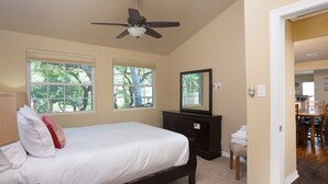 Ein Schlafzimmer mit einem Bett, Deckenventilator, Kommode und Spiegel. Fenster bieten Blick auf Bäume.