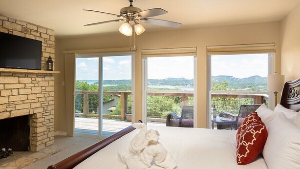 Ein Schlafzimmer mit Kamin aus Stein, einem Bett mit weißer Bettwäsche, einem Deckenventilator und großen Fenstern mit Blick ins Freie.