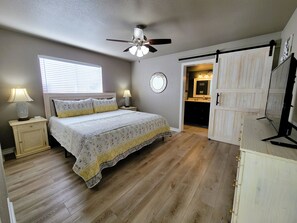 A bedroom with a bed, a ceiling fan, a window with blinds, a nightstand with a lamp, and a door leading to a bathroom. - Shabby Chic Remodel by the Lake! (Canyon Lake)