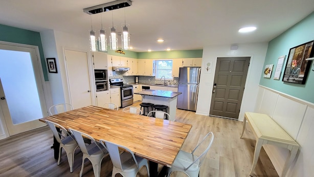 Una cocina moderna con una mesa de comedor de madera, sillas blancas, electrodomésticos de acero inoxidable e iluminación colgante.