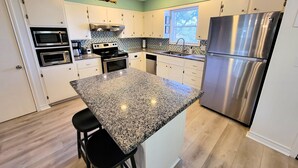 Una cocina moderna con armarios blancos, encimeras de granito, electrodomésticos de acero inoxidable y un ventanal que deja entrar luz natural.