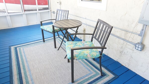 Un espace patio doté d’un sol bleu, d’une table ronde, de deux chaises avec des coussins à motifs floraux et d’une boîte de prises électriques fixée au mur.