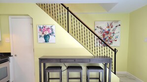 A kitchen with a dark wooden table and two stools, a staircase with a black railing, and two floral paintings on the walls. - 2nd-Story Covered Deck with Lake View - 2 Kings, 3 Queens! (Canyon Lake)