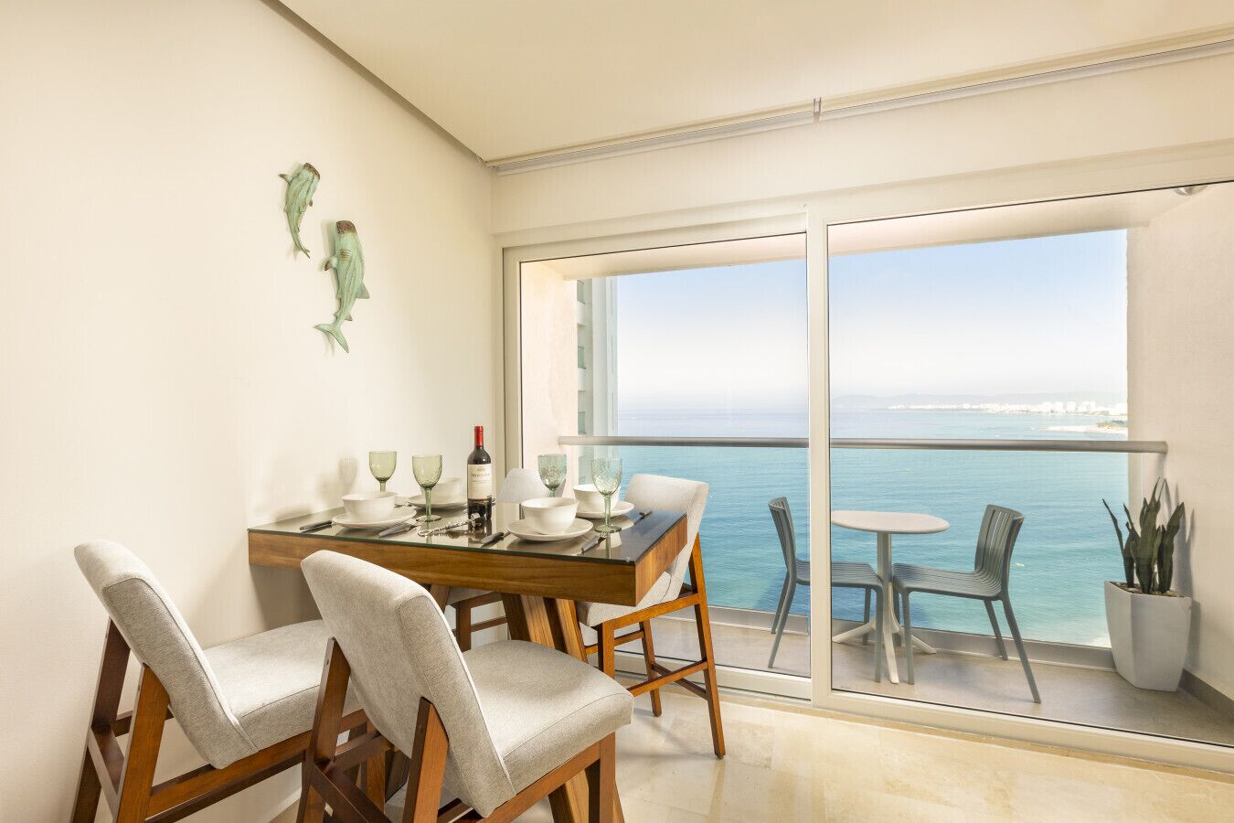 A dining area with a table set for four, a view of the sea through large windows, and a balcony with chairs.