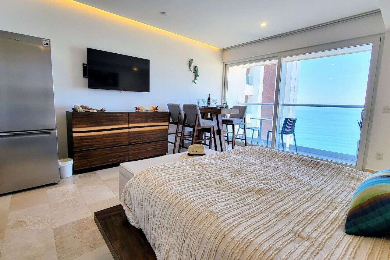 Ein modernes Schlafzimmer mit einem großen Bett, einem Flachbildfernseher, einem Essbereich mit Stühlen und Meerblick durch große Fenster.