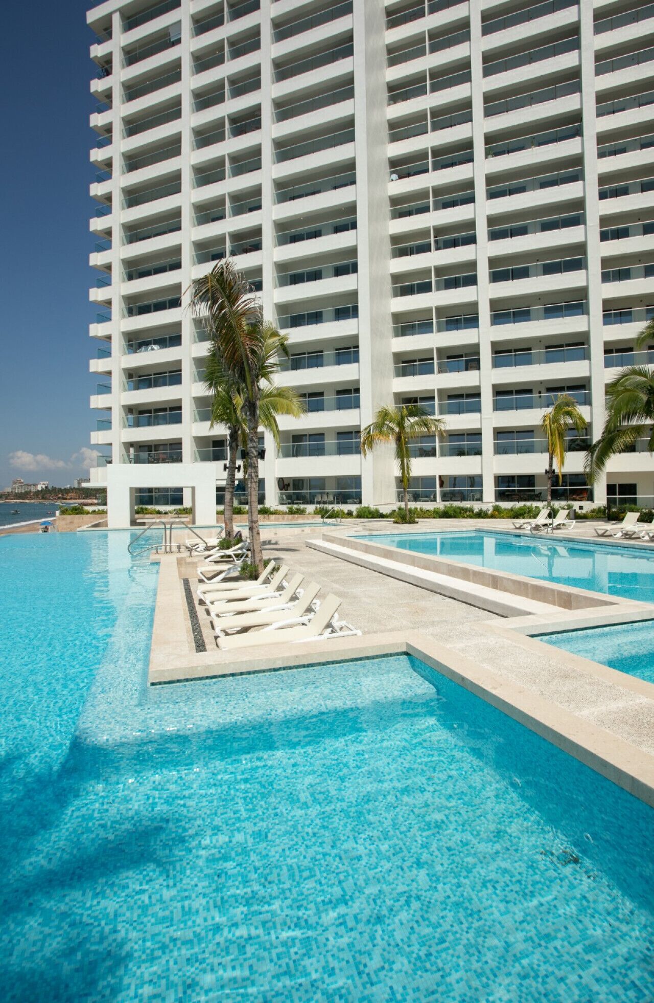 Un hôtel moderne doté d’une grande piscine, de nombreux fauteuils de relaxation et de palmiers.