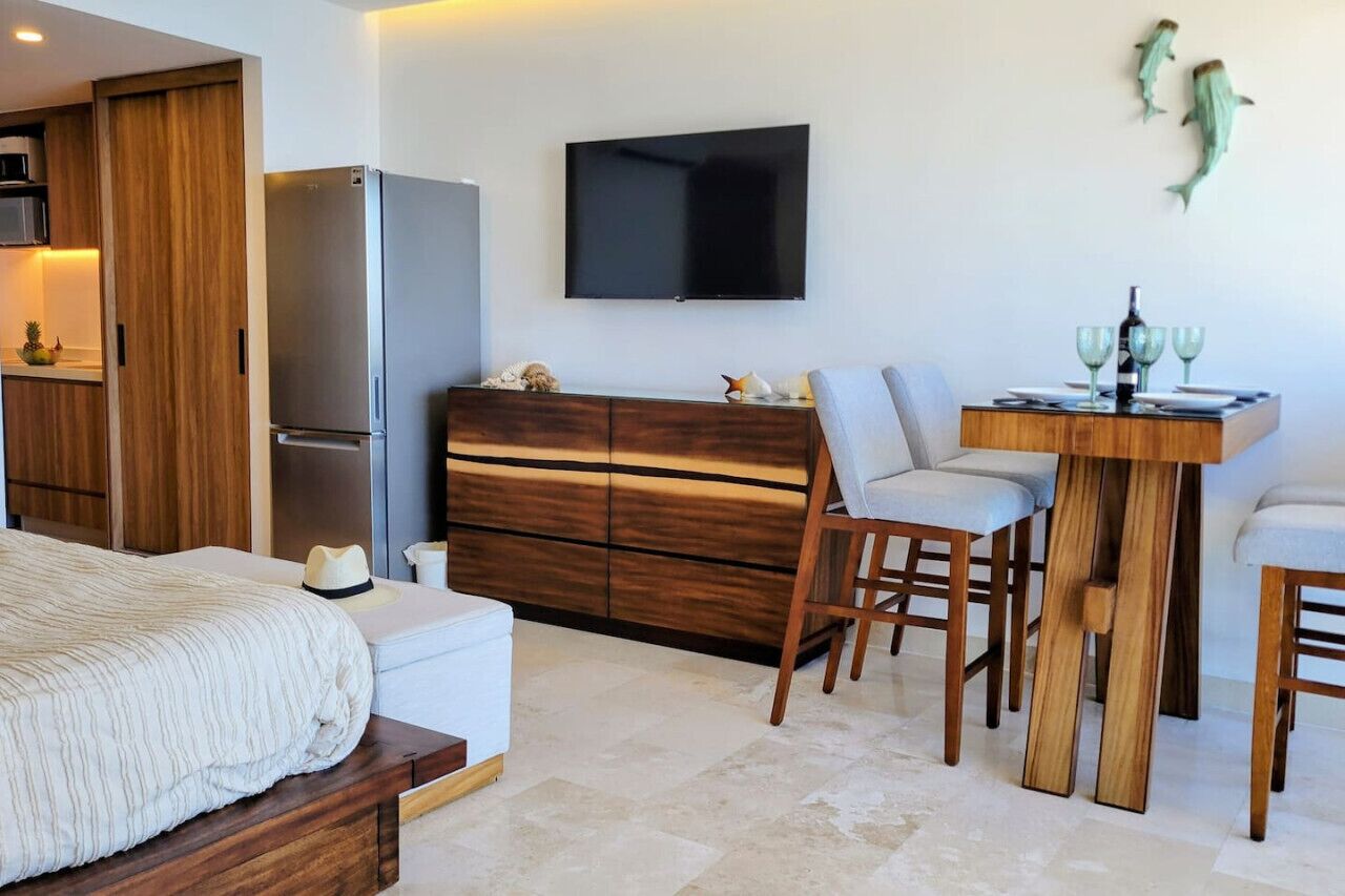 A modern hotel room with a bed, a wooden dresser, a TV mounted on the wall, and a dining area with a table and chairs.