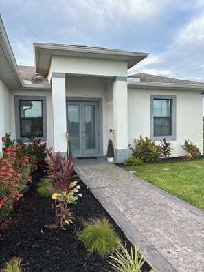A residential building with a front entrance, a covered porch, and a landscaped garden with various plants and flowers. - Belle Ville  (Lehigh Acres)