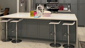 A modern kitchen with grey cabinets, a stainless steel refrigerator, a microwave, and a gas stove. There are two bar stools and a large island with a white countertop. - Belle Ville  (Lehigh Acres)