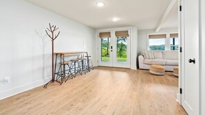 A spacious room with a wooden floor, a dining area with a table and chairs, a sofa, and a view of greenery through the door.