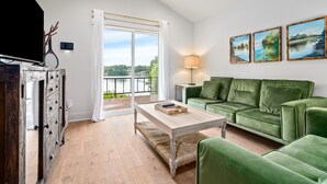 A living room with a green sofa, a wooden coffee table, and a TV on a wooden cabinet. There is a window with a view of greenery and a lamp on a side table.
