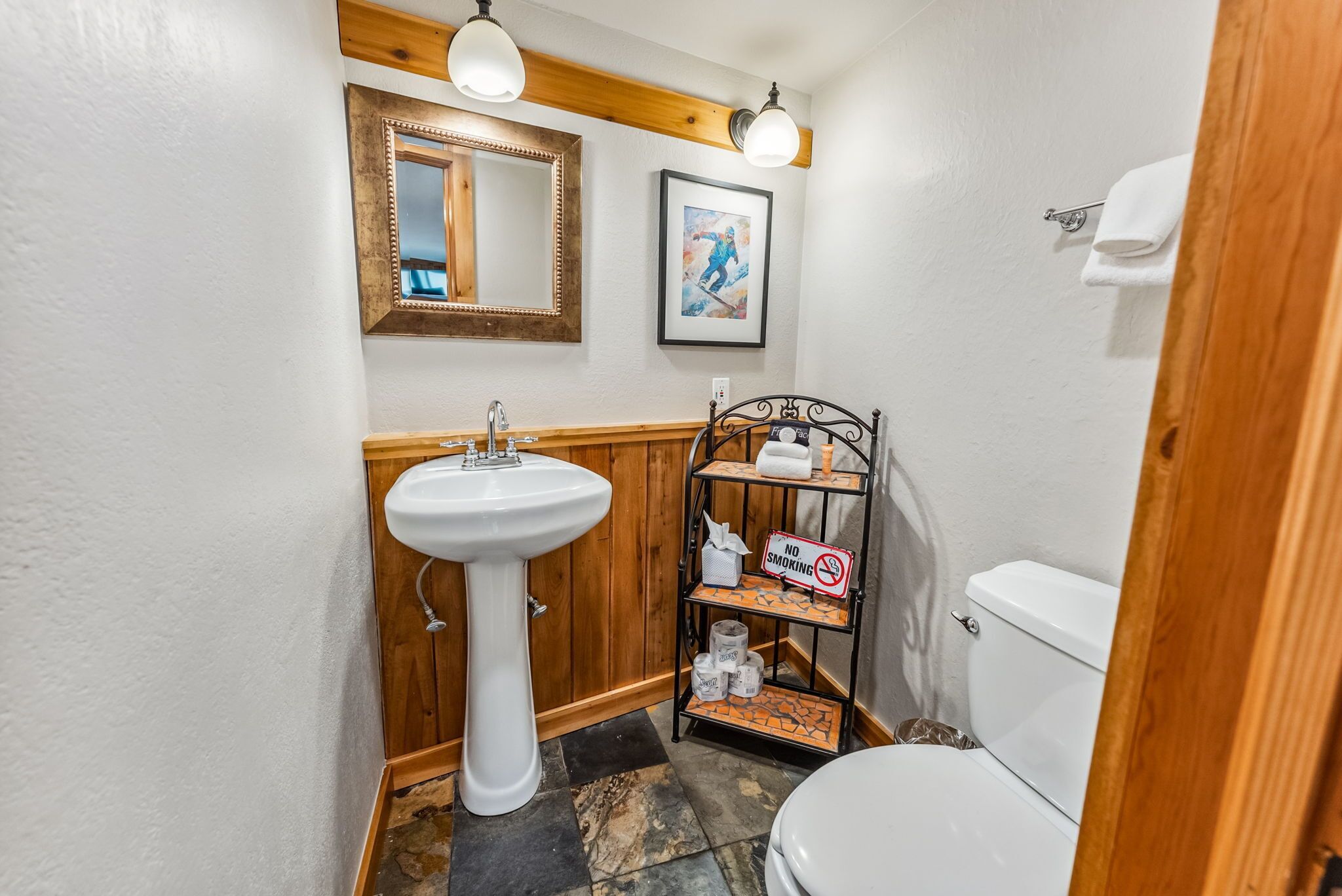 Een badkamer met een wastafel op een voetstuk, een houten wastafelmeubel, een lijst met spiegel, een bordje ‘Niet roken’, een plank met toiletartikelen en een toilet.