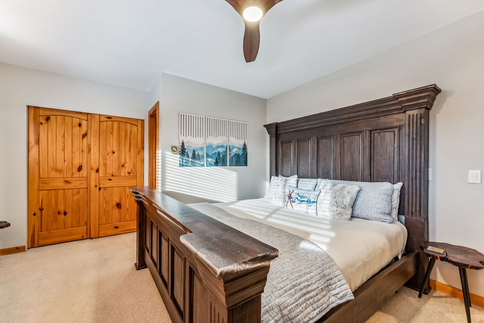 A bedroom with a wooden headboard, a large bed with multiple pillows, a wooden bench, a ceiling fan, and a window with blinds.