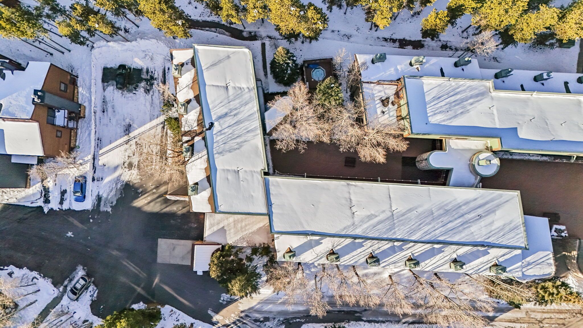 Vista aérea de uma área residencial com vários edifícios, incluindo uma casa com garagem visível e uma garagem separada. Árvores e um pequeno lago são visíveis.
