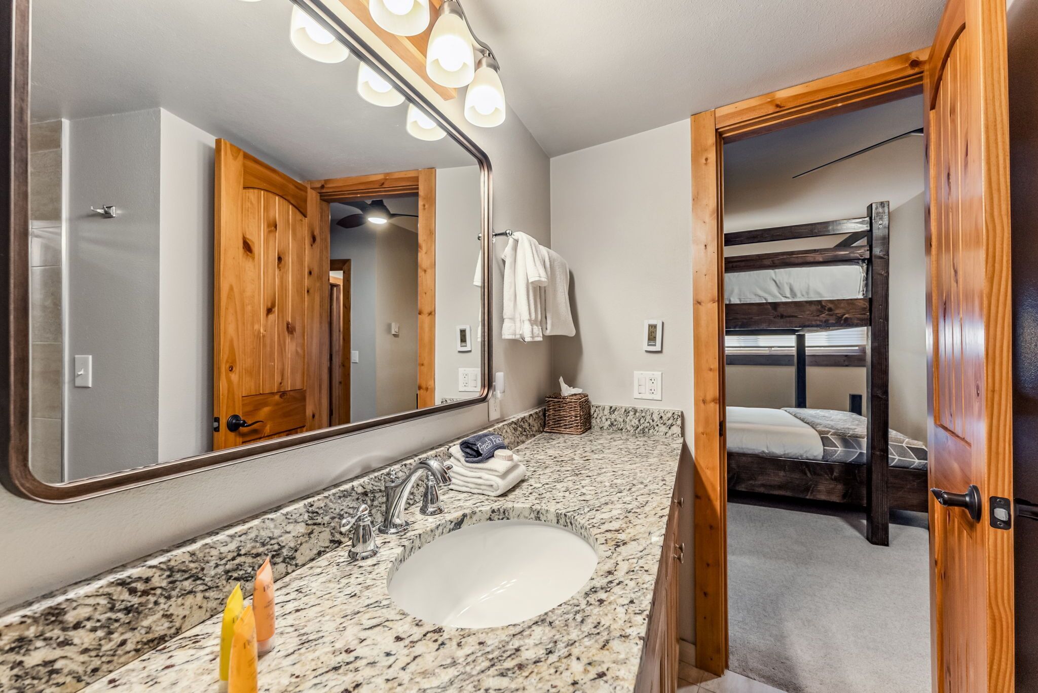 A bathroom with a granite countertop, a sink, and a mirror with lights. There is a wooden door leading to a bunk bed.