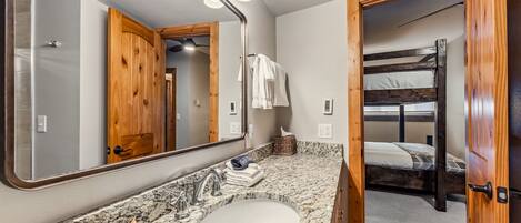 A bathroom with a granite countertop, a sink, and a mirror with lights. There is a wooden door leading to a bunk bed.