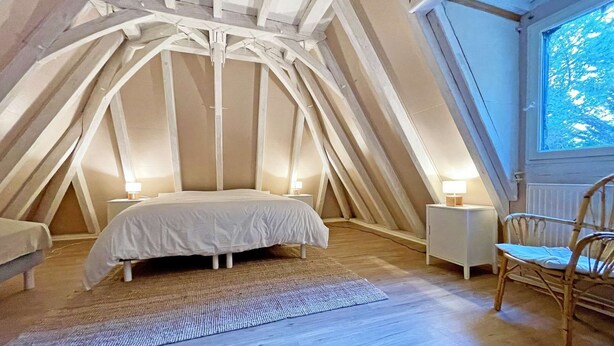A bedroom with a sloped ceiling, a bed, a chair, and a window.