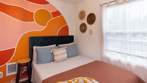 A bedroom with a bed, a stool, a wall mural, and a window with blinds.