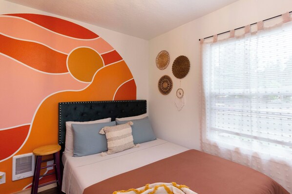Une chambre à coucher comprenant un lit, un tabouret, une fresque murale et une fenêtre avec des stores.
