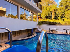 Una piscina con un tobogán azul, un espacio exterior iluminado por el sol con una mesa y sillas, y un edificio con grandes ventanales.