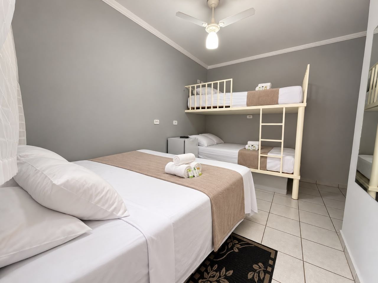 Een hotelkamer met een eenpersoonsbed, een stapelbed, een plafondventilator en een nachtkastje met een lamp.