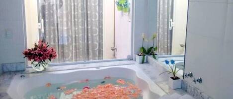 Un baño con una bañera grande llena de agua y pétalos de rosa, un espejo y una ventana con cortinas.