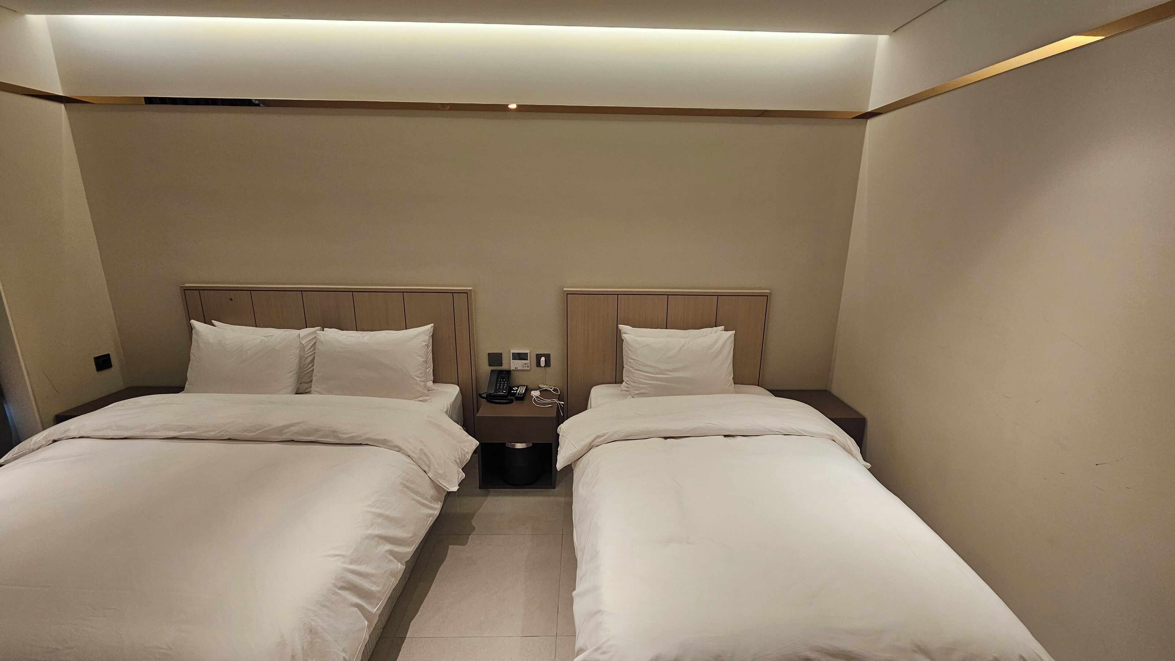 Quarto de hotel com duas camas de solteiro, uma mesa de cabeceira com telefone e luminária, e um criado-mudo pequeno.