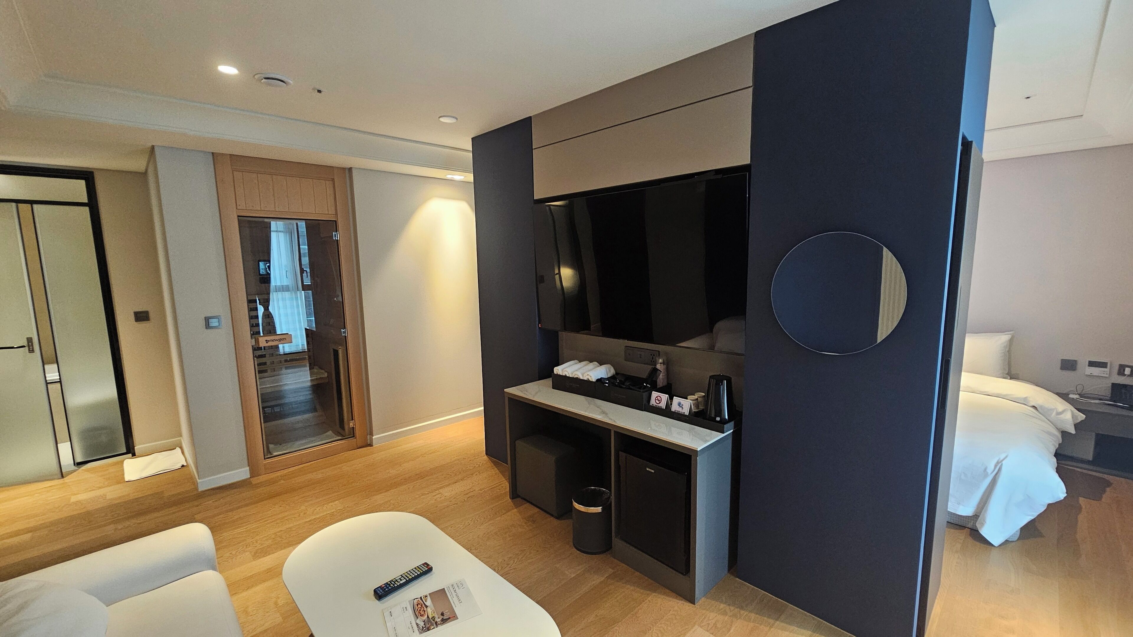 A modern hotel room with a large bed, a desk with a flat-screen TV, a seating area with a white chair, and a small trash bin.