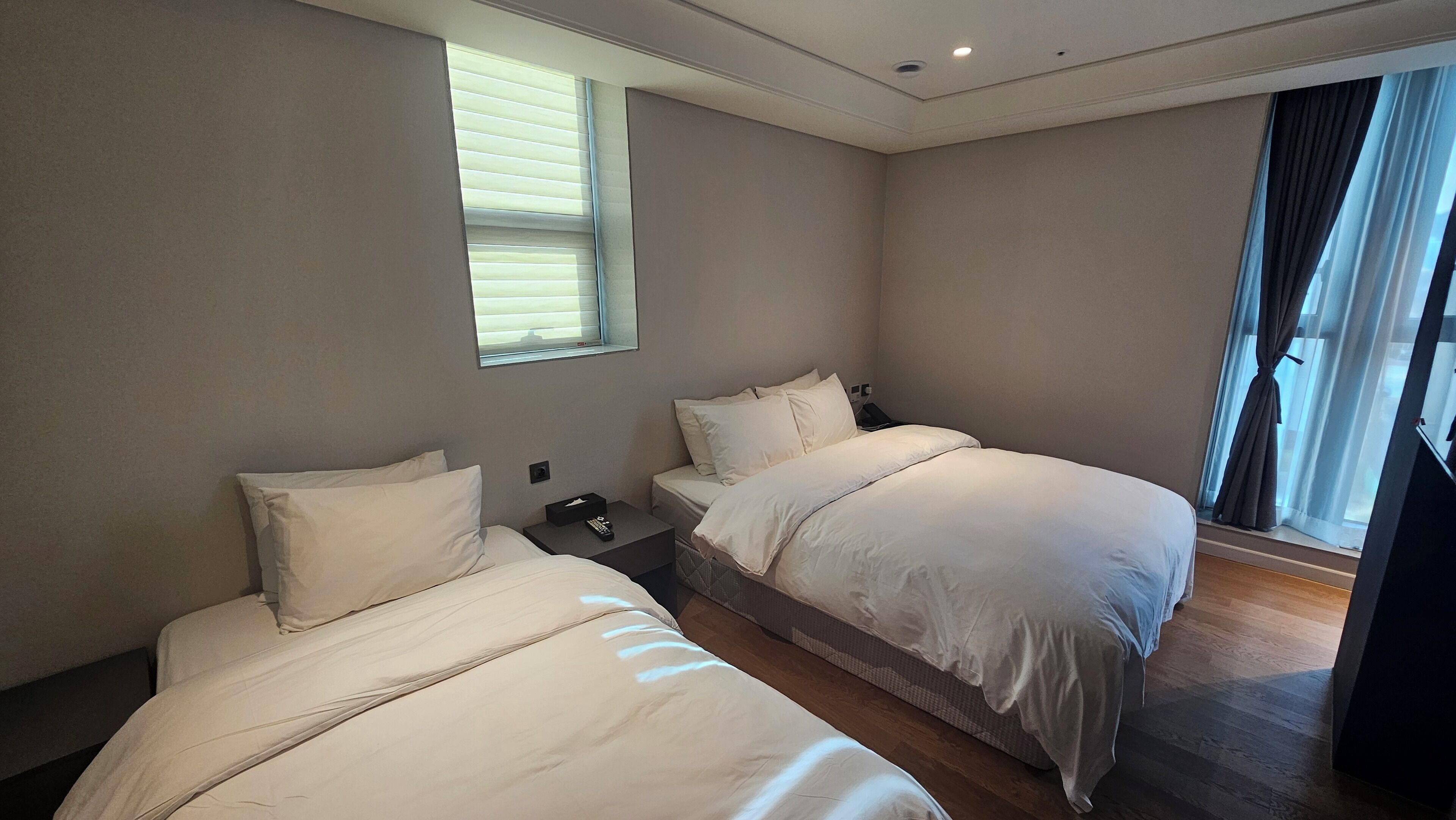 A hotel room with two beds, a window with blinds, a nightstand, and a view of the outside.