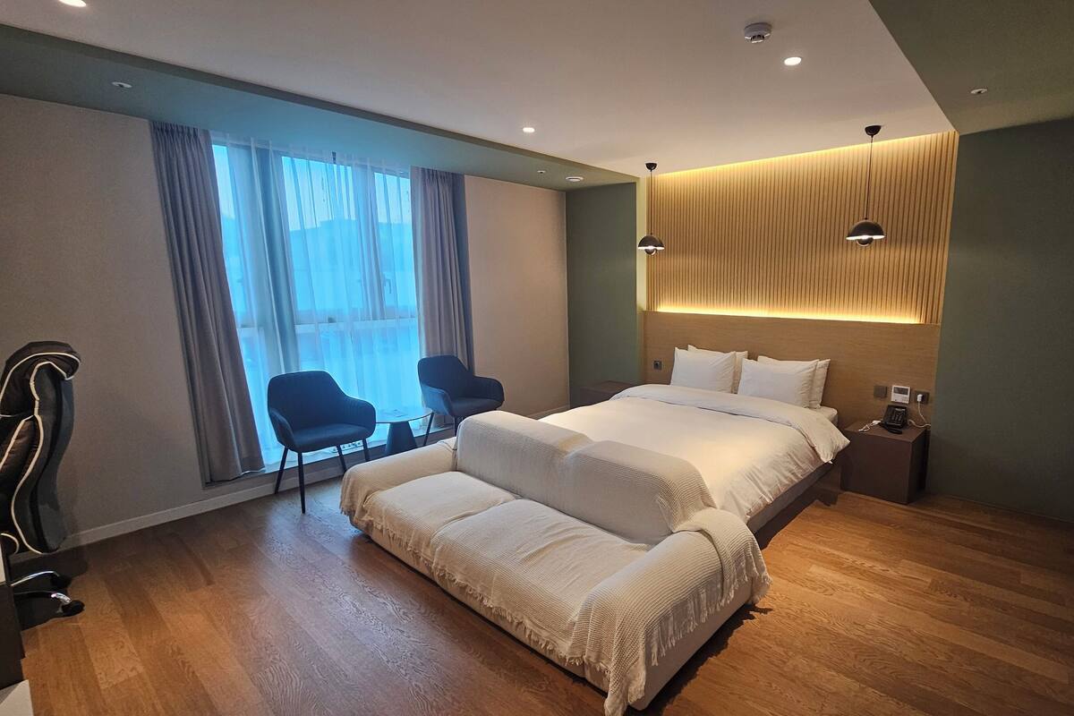 Quarto de hotel moderno com uma cama grande, um sofá, duas cadeiras, uma mesinha de cabeceira com telefone e uma janela com cortinas.