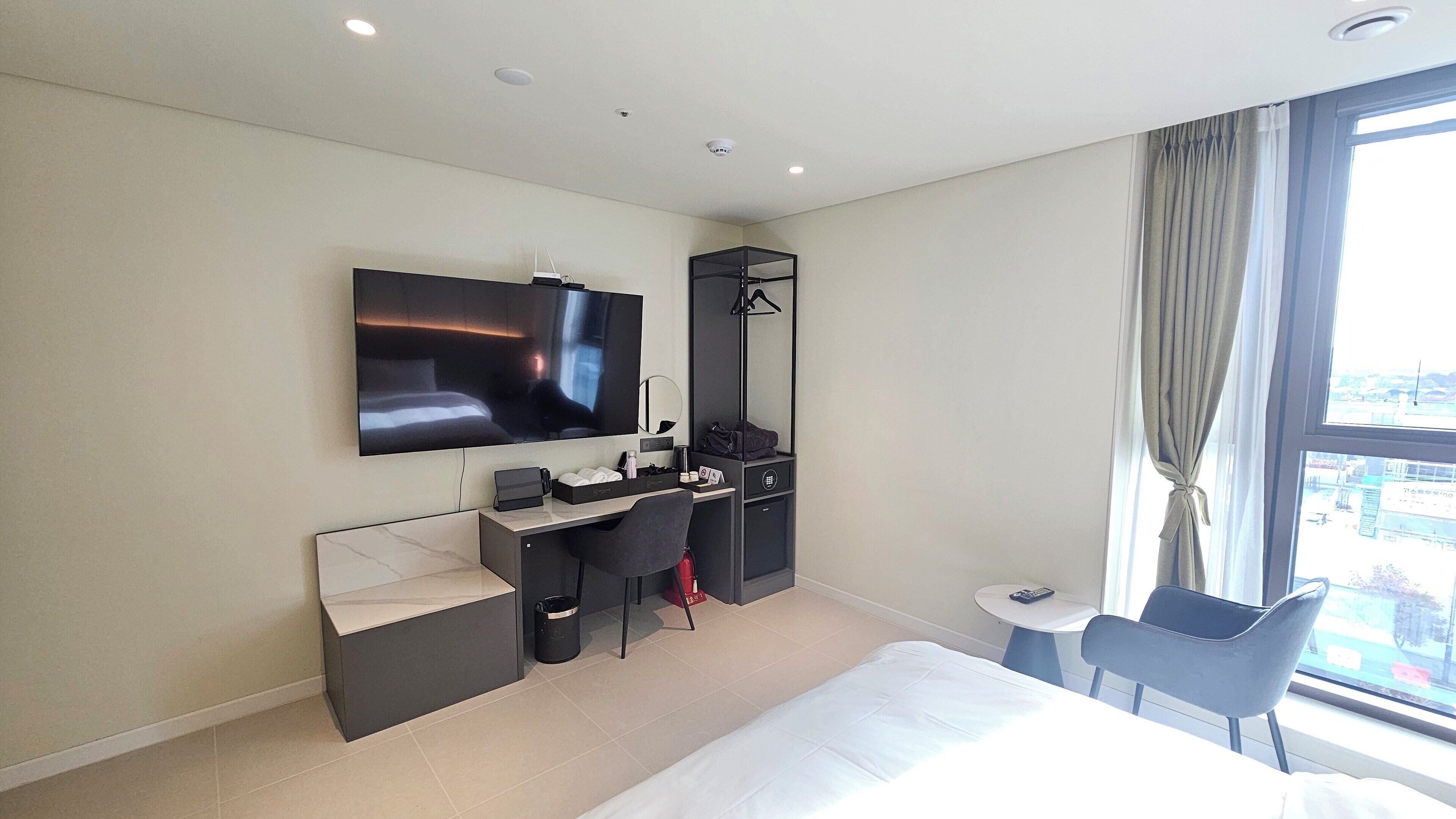 A modern hotel room with a large bed, a desk with a chair, a TV mounted on the wall, a small table, and a window with curtains.