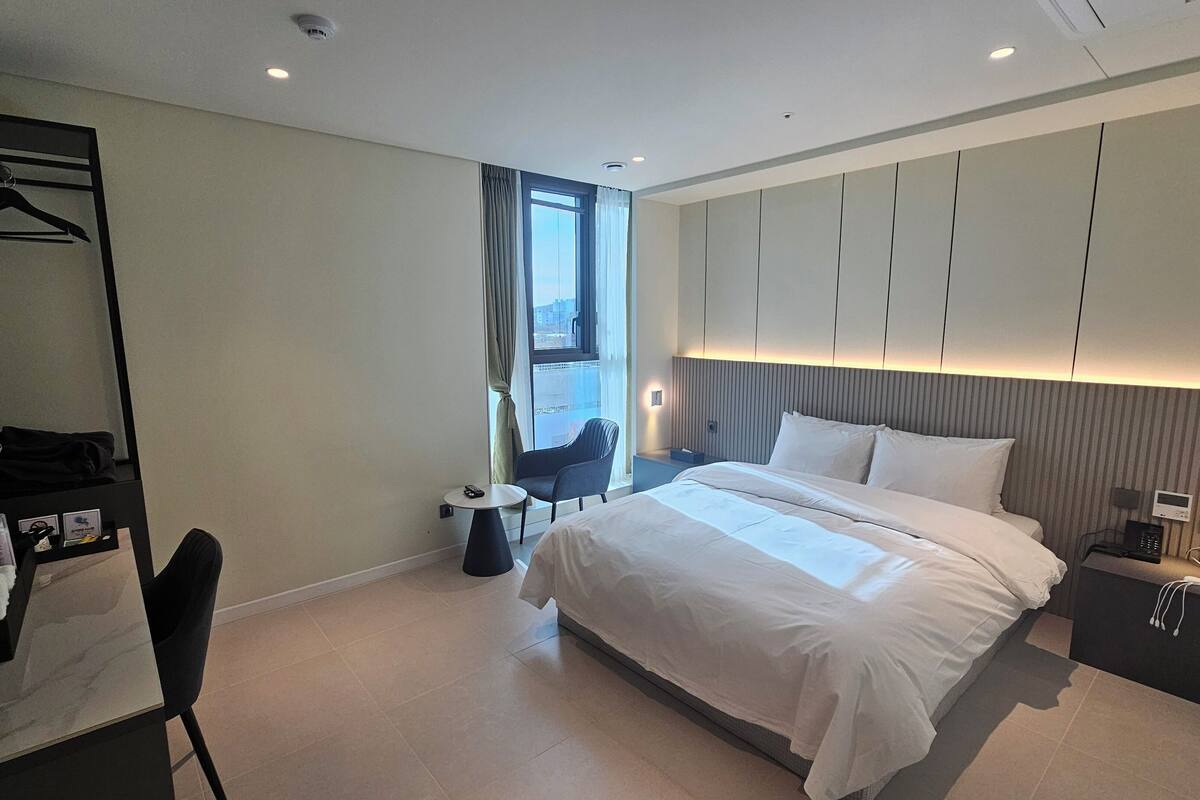 Quarto de hotel moderno com uma cama grande, uma escrivaninha com cadeira, um guarda-roupa e uma pequena mesa.