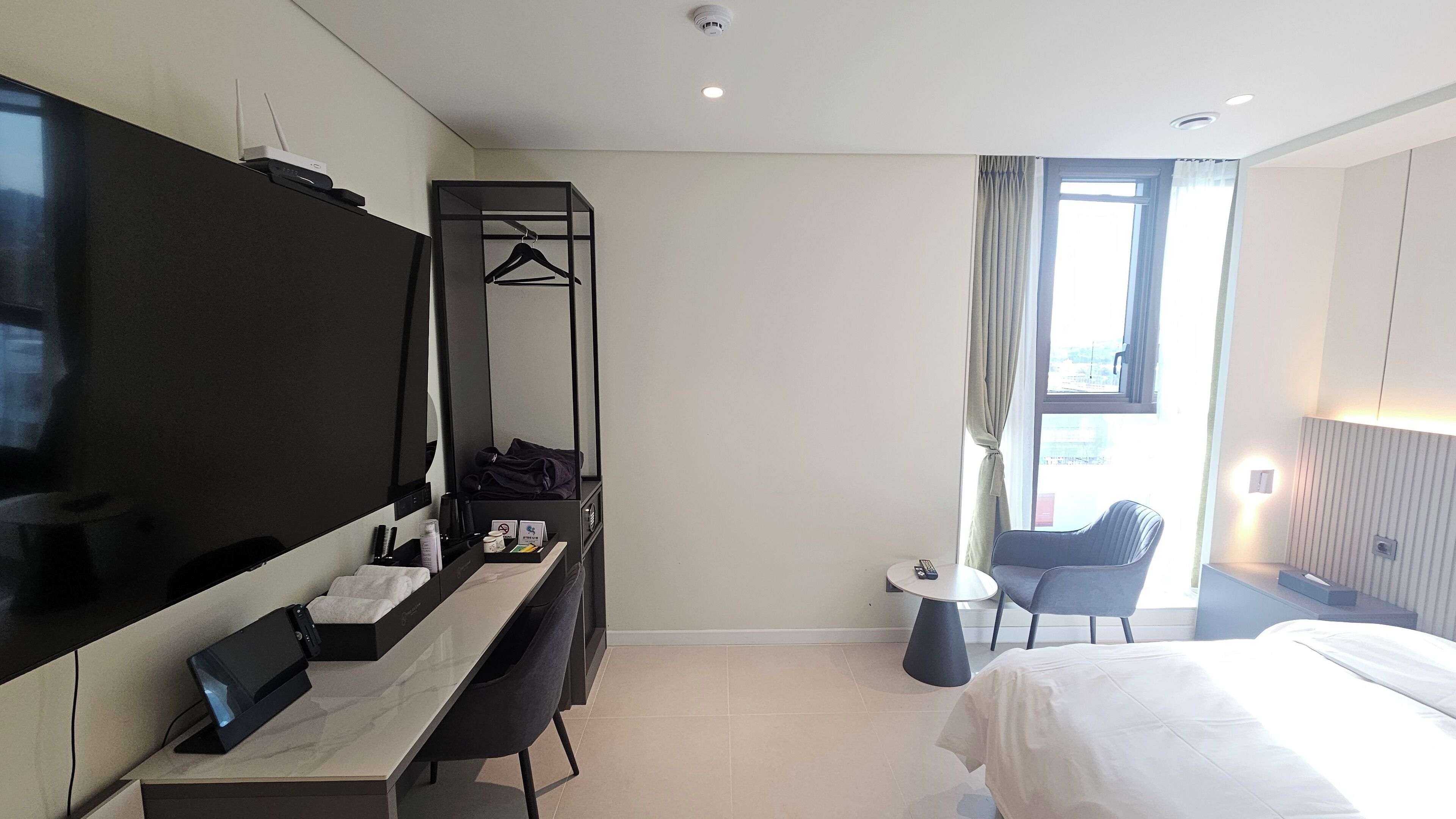 A modern hotel room with a flat-screen TV, a desk with a chair, a bed, a small table with a chair, and a window with curtains.