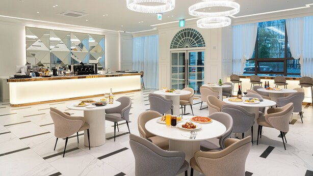 A modern dining area with round tables and chairs, a central bar, and large windows with curtains. - The Gleam Hotel (Incheon)