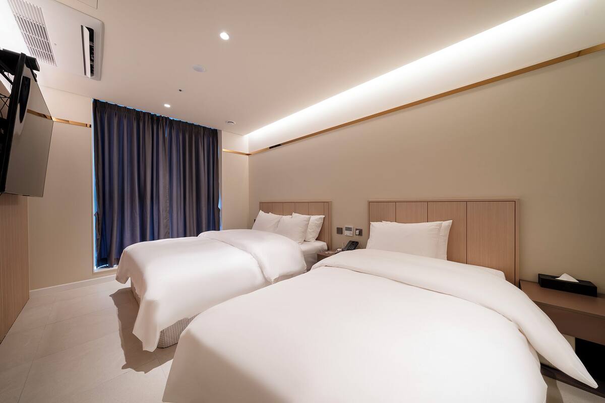 Quarto de hotel com duas camas, televisão, escrivaninha e janela com cortinas.